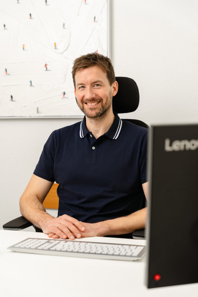 Tim Grüber sitzt lächelnd an seinem Schreibtisch und schaut in die Kamera. Im Hintergrund Kunstwerk mit Figuren.
