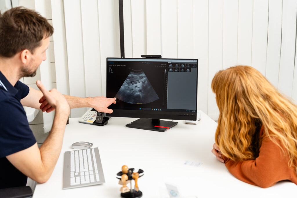 Tim Grüber erklärt einer Patientin ein Ultraschallbild des Pankreas auf einem Monitor in einer Arztpraxis. Medizinische Untersuchung und Diagnose.